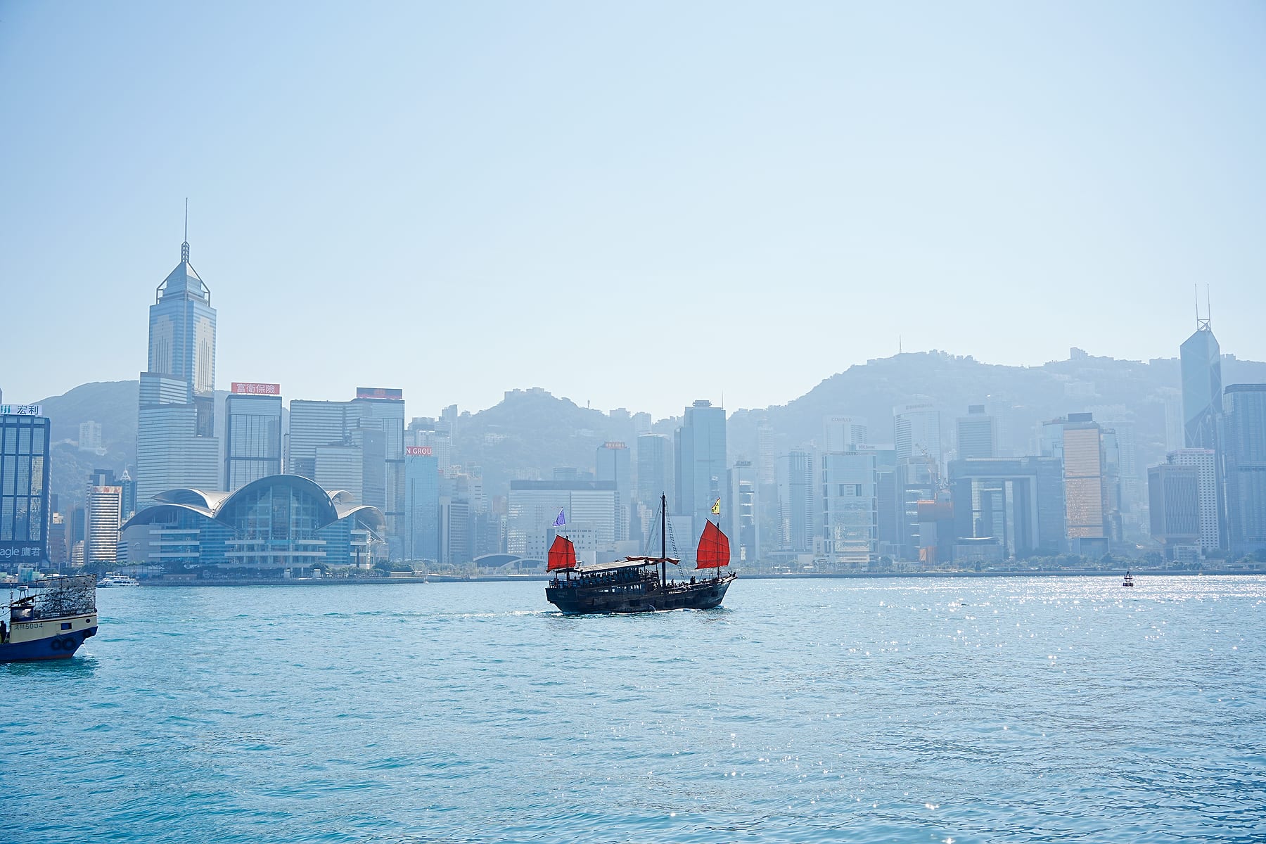 蓝色海港中，一艘红帆船驶过城市与山影