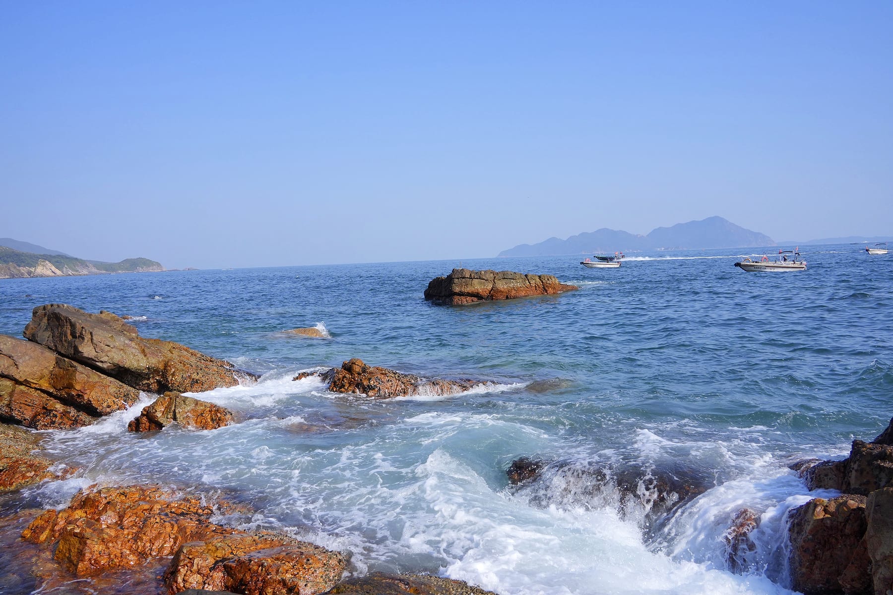 海浪拍向褐色礁石，远处小船散在蓝色海面