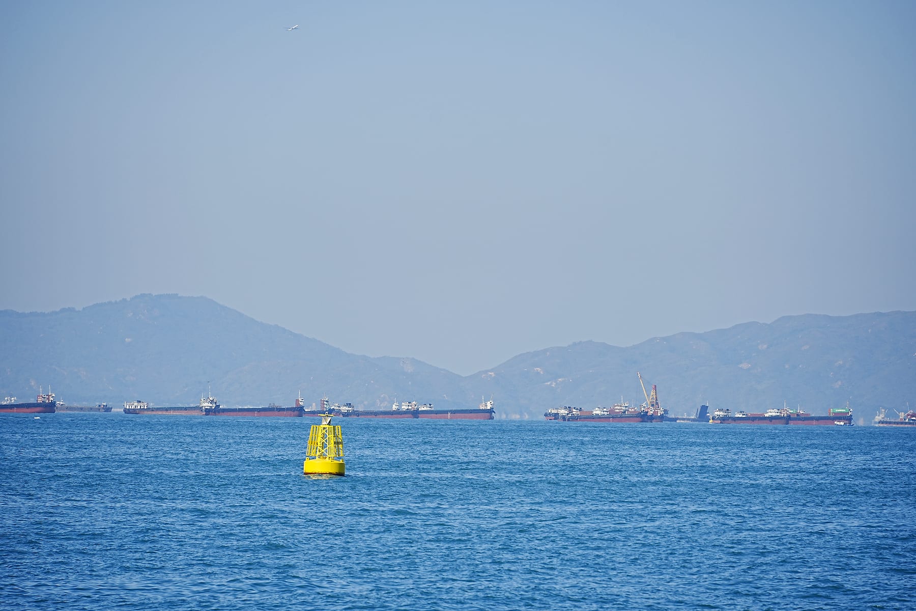 蓝色海面上一枚黄色浮标,远处货船沿山脚排开
